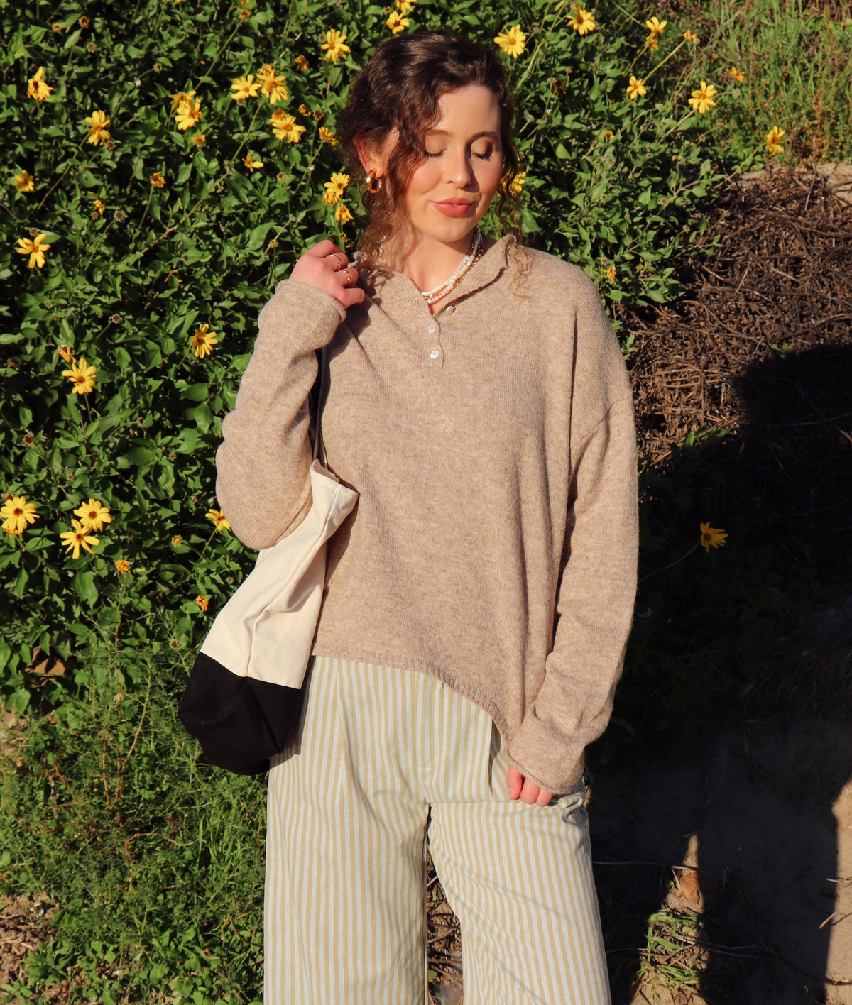 Woman wearing a beige sweater top and striped pants standing in front of greenery with yellow flowers.