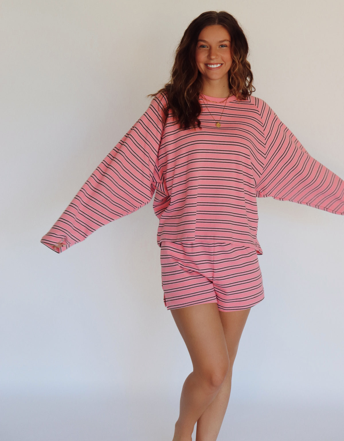 A woman modeling a pink and brown striped lounge set consisting of a long-sleeve top and matching shorts, posed against a plain white studio background.