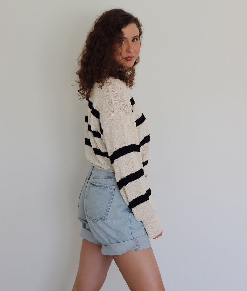 A woman with curly brown hair wearing a cream-colored knit sweater with bold black horizontal stripes and light-wash denim shorts, standing against a plain light gray wall.