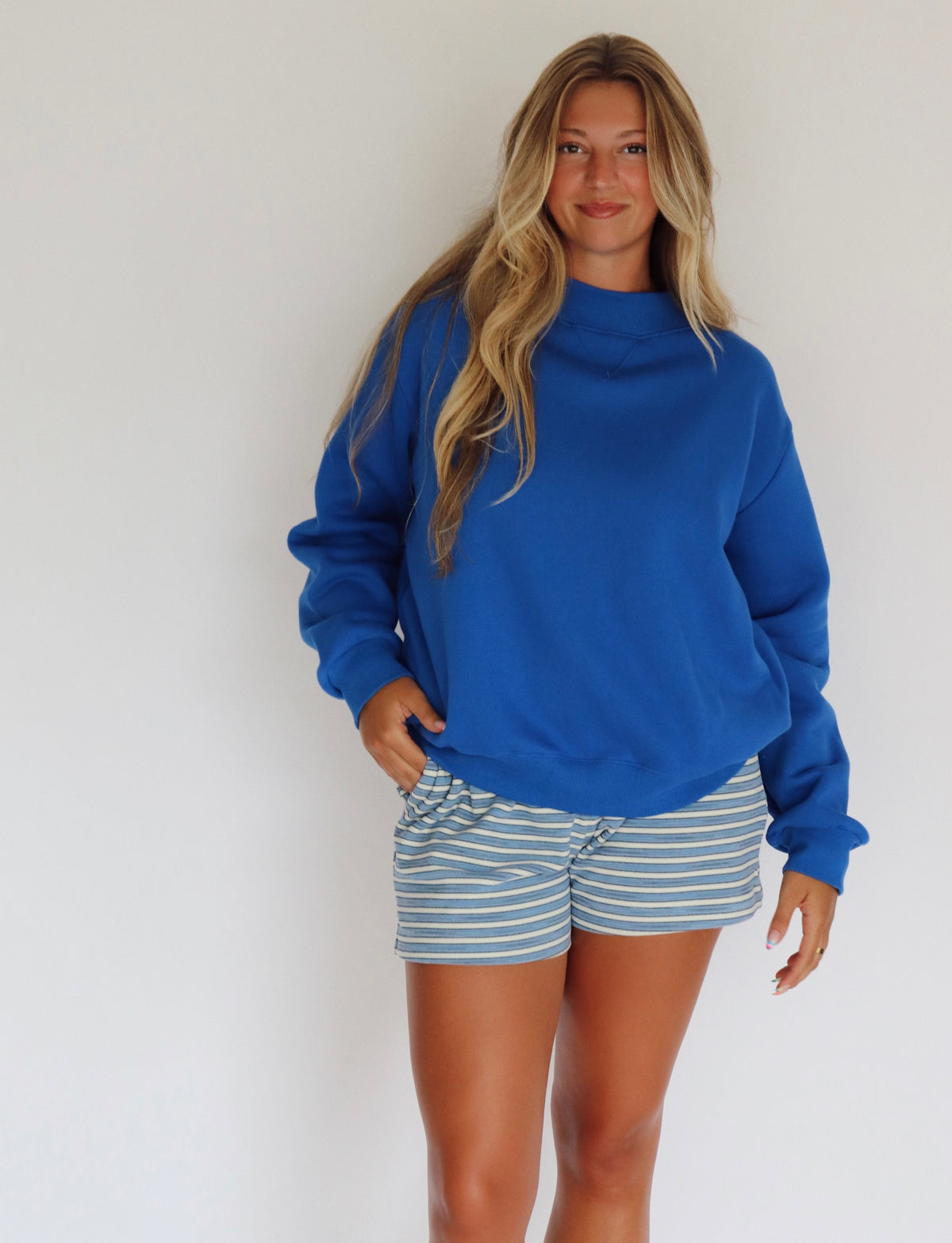 A woman modeling a royal blue mock neck sweatshirt and blue and white striped shorts against a plain white wall.