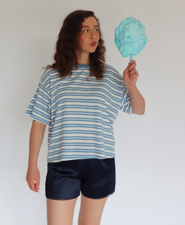 A woman with curly brown hair stands against a plain white background, wearing a loose blue and white striped t-shirt and dark navy shorts, while holding a light blue cotton candy stick.