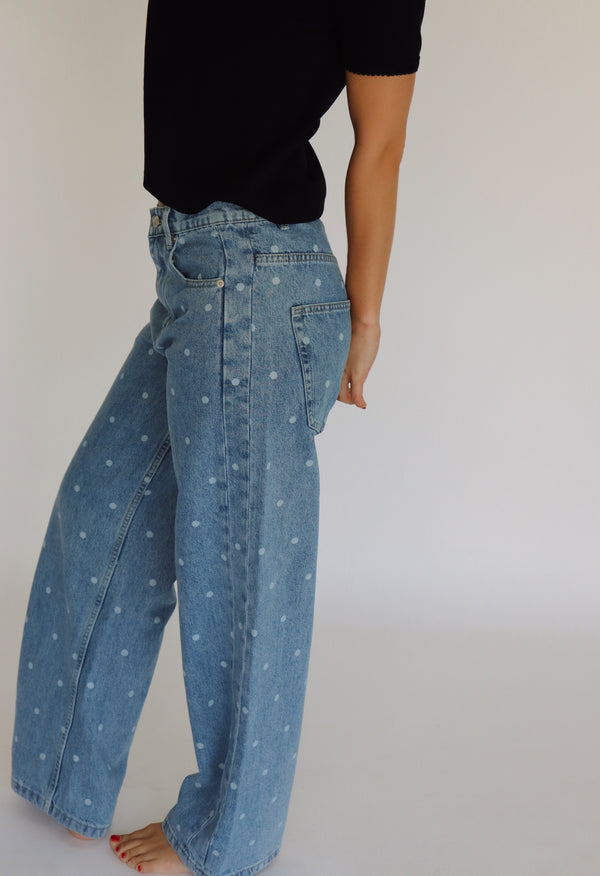A woman modeling a black short-sleeve top and light blue wide-leg denim jeans with small white polka dots, standing against a plain white studio background.