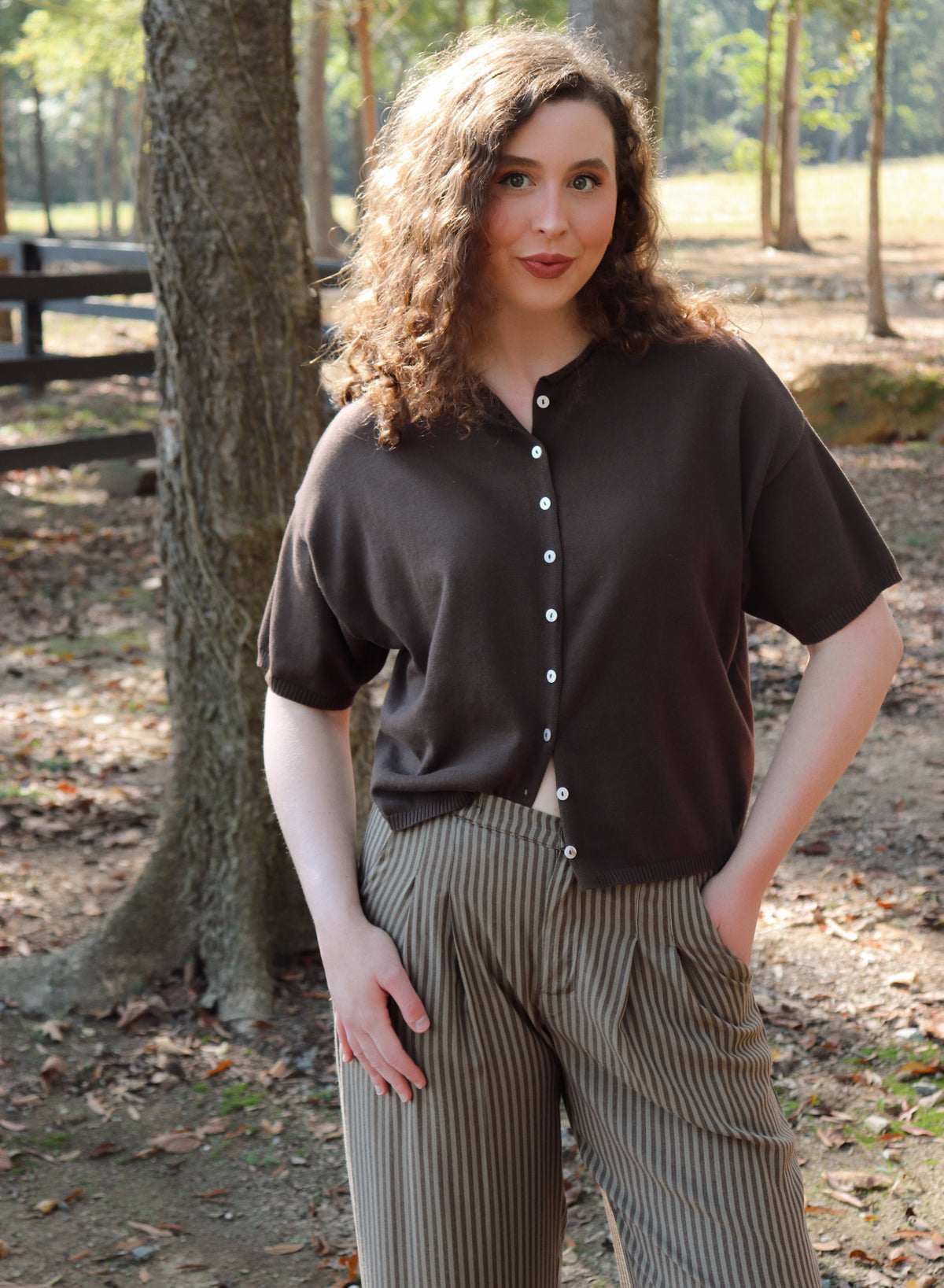 a woman wearing a chocolate brown short sleeve cardigan with brown striped pants stands in an outdoor setting