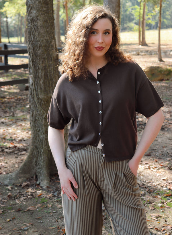a woman wearing a chocolate brown short sleeve cardigan with brown striped pants stands in an outdoor setting