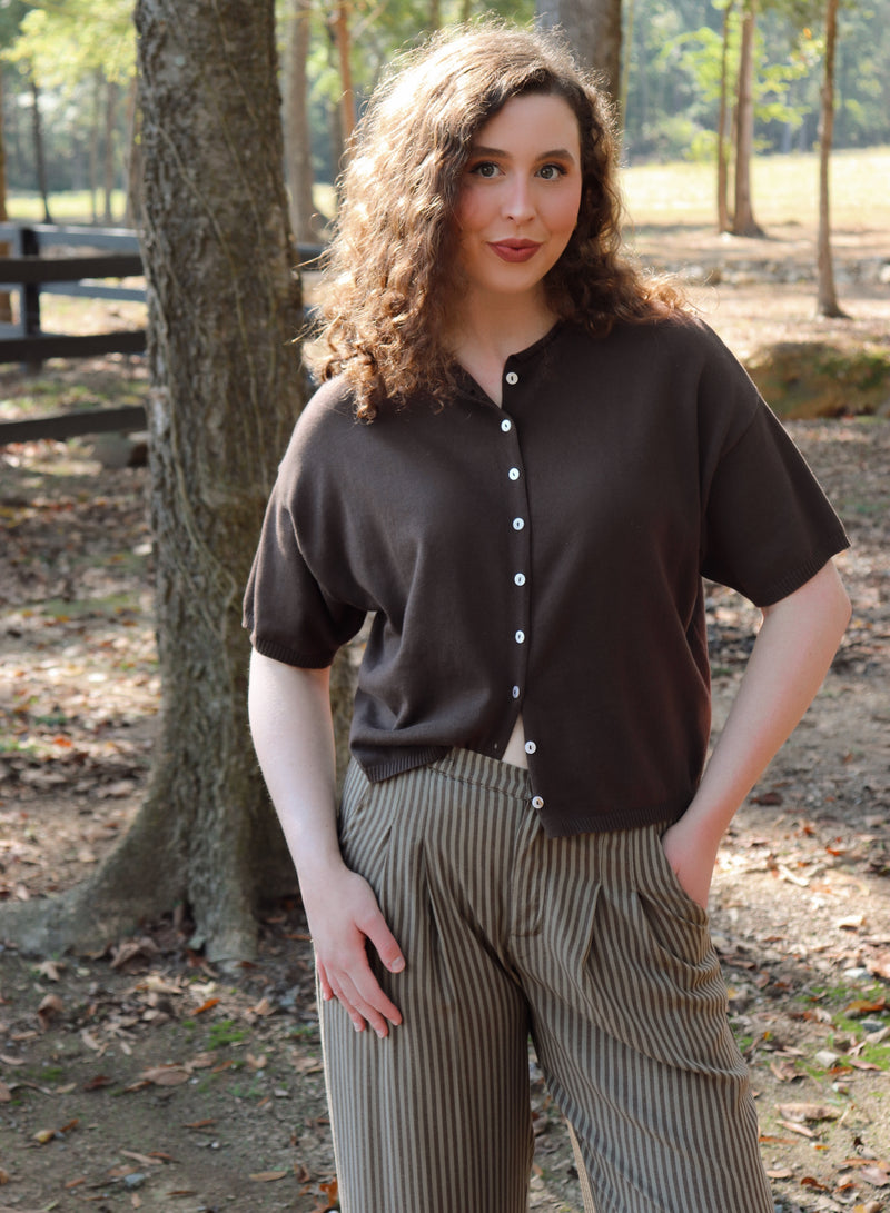 a woman wearing a chocolate brown short sleeve cardigan with brown striped pants stands in an outdoor setting