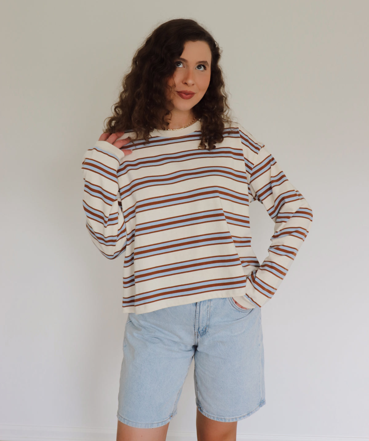 A woman with curly brown hair wearing a cream-colored long-sleeve knit sweater with thin blue and camel horizontal stripes, paired with light blue denim shorts, standing against a plain off-white wall.