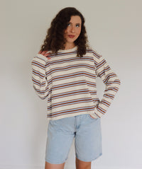 A woman with curly brown hair wearing a cream-colored long-sleeve knit sweater with thin blue and camel horizontal stripes, paired with light blue denim shorts, standing against a plain off-white wall.