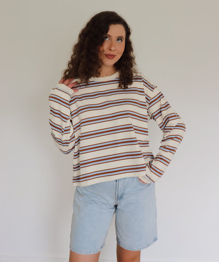 A woman with curly brown hair wearing a cream-colored long-sleeve knit sweater with thin blue and camel horizontal stripes, paired with light blue denim shorts, standing against a plain off-white wall.
