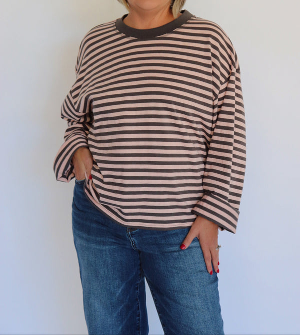 A woman wearing a brown and gray striped long-sleeve top and blue jeans, standing against a plain white wall.