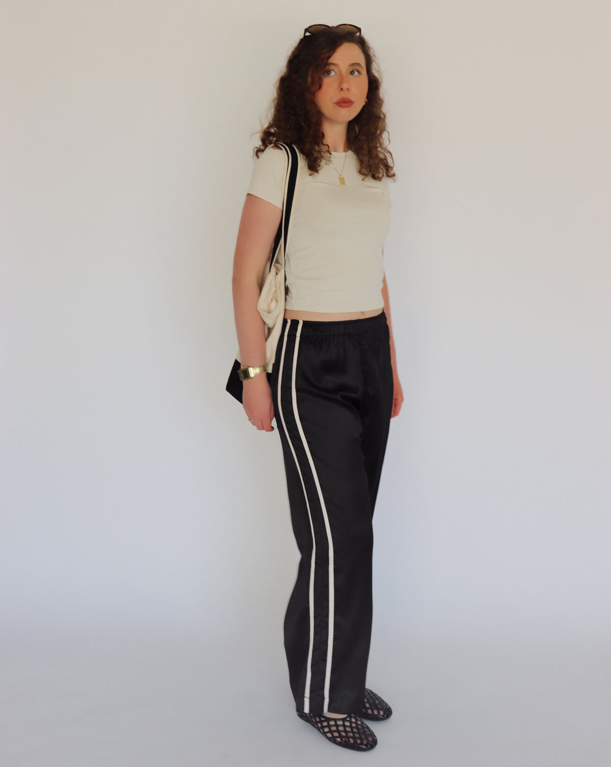 A woman stands against a plain light background wearing a cream crop top, black wide-leg pants with a white side stripe, black  flats, and carrying a black and cream tote bag.