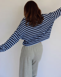 A woman wearing a navy and light blue horizontally striped long-sleeve cardigan and light gray sweatpants, standing against a plain off-white wall.