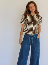 A woman wearing a brown and beige vertically striped short-sleeve button-up top with blue wide-leg jeans, standing against a plain light gray wall.