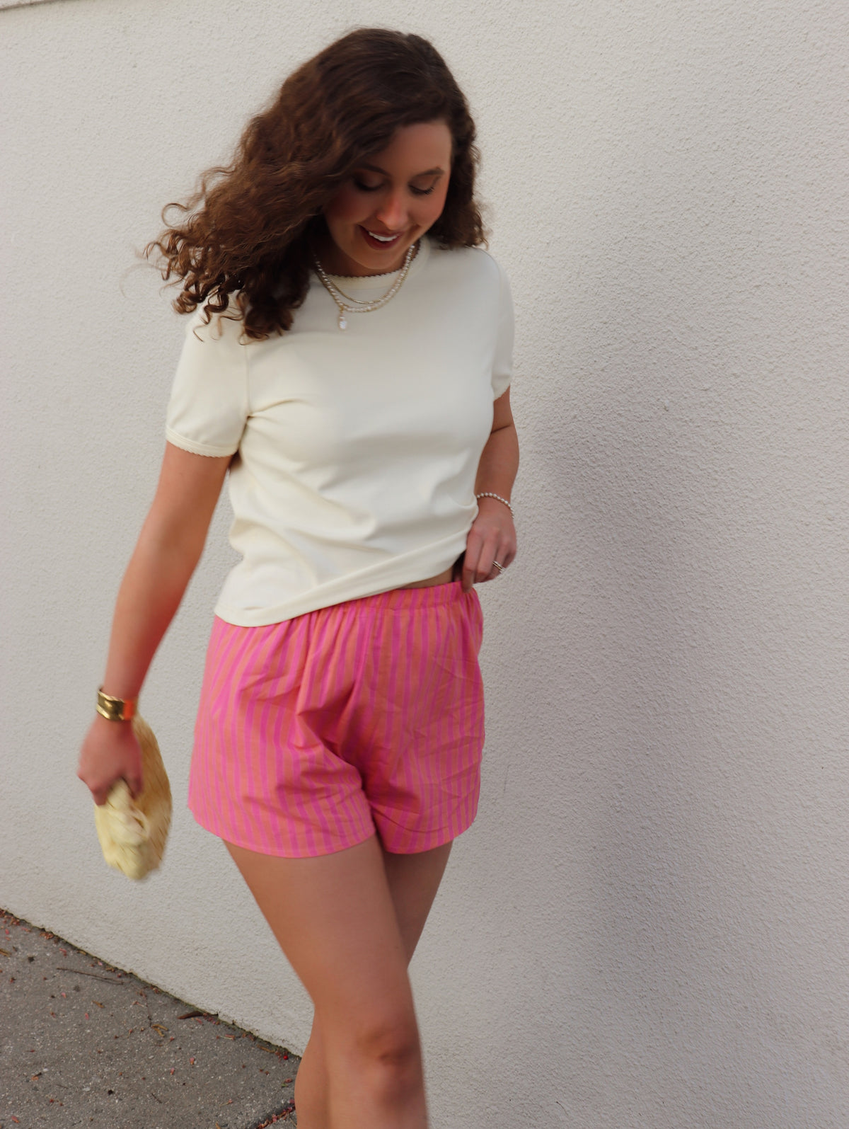 Woman wearing a cream shirt and orange and fuchsia striped shorts against a light gray wall.