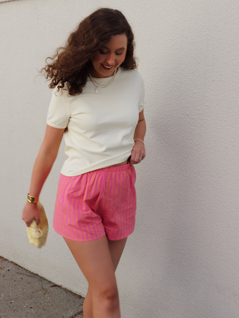 Woman wearing a cream shirt and orange and fuchsia striped shorts against a light gray wall.