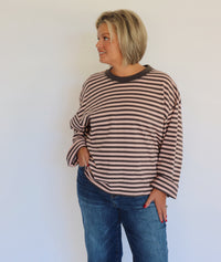 A woman with a short blonde bob wearing a brown and gray striped long-sleeve top and blue jeans, standing against a plain white wall.