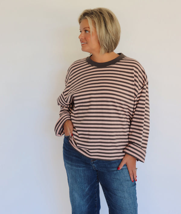A woman with a short blonde bob wearing a brown and gray striped long-sleeve top and blue jeans, standing against a plain white wall.