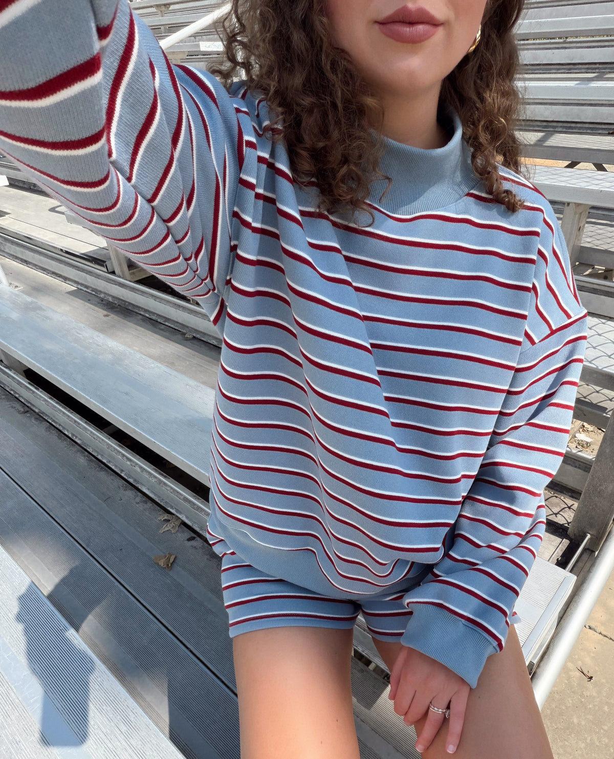 A person wearing a light blue long-sleeve sweatshirt with thin red and white horizontal stripes, paired with matching striped shorts, sitting on outdoor metal bleachers.