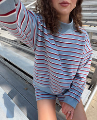 A person wearing a light blue long-sleeve sweatshirt with thin red and white horizontal stripes, paired with matching striped shorts, sitting on outdoor metal bleachers.