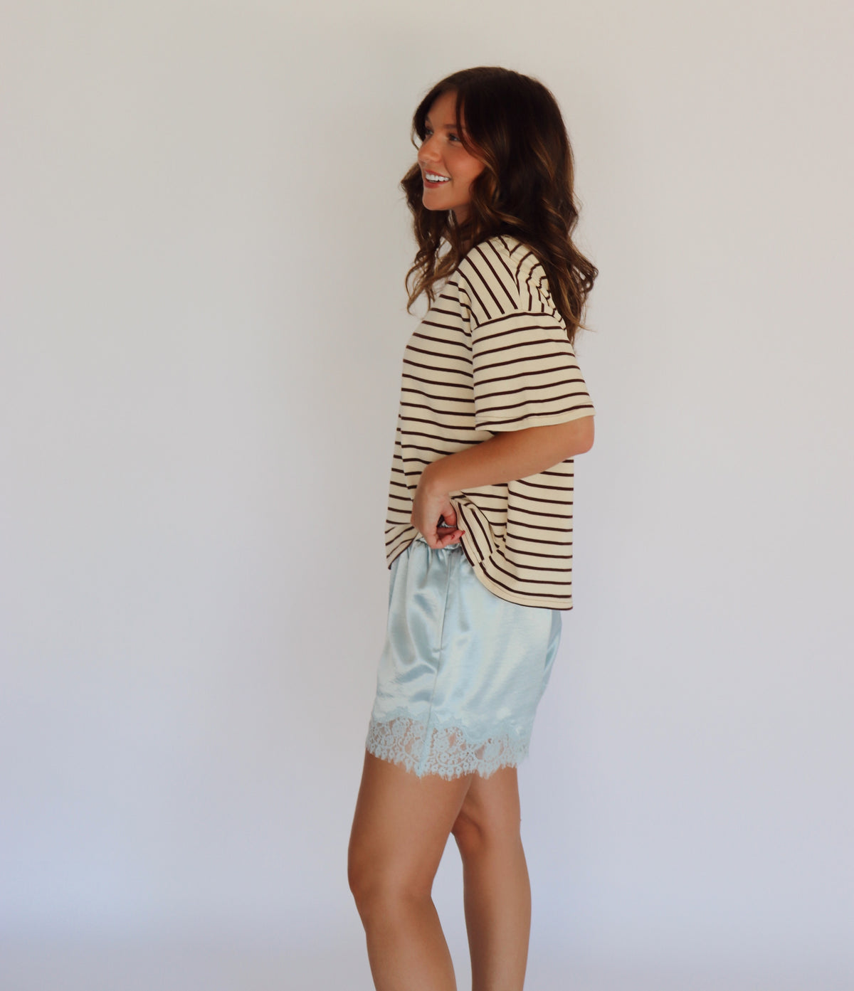 A woman modeling a beige and brown horizontally striped short-sleeve t-shirt with light blue satin shorts featuring a lace hem, standing against a plain white studio background.