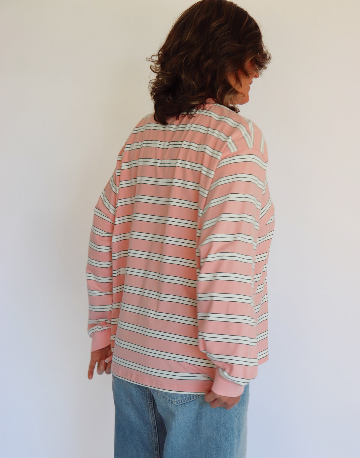 A woman wearing a pink long-sleeve crewneck shirt with thin white horizontal stripes, paired with light blue jeans, standing against a plain off-white wall.