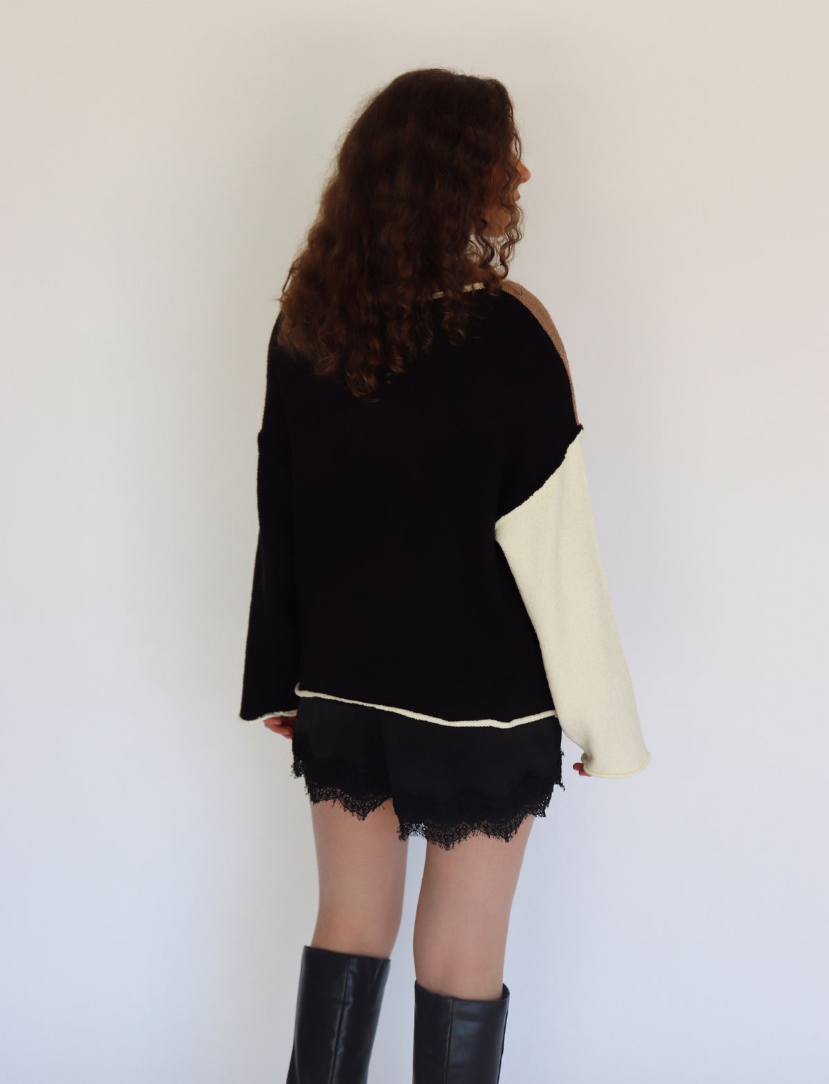A woman shown from the back wearing a black knit sweater with one contrasting cream-colored sleeve, black lace-trimmed shorts, and black knee-high boots, set against a plain white wall.