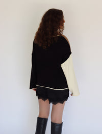 A woman shown from the back wearing a black knit sweater with one contrasting cream-colored sleeve, black lace-trimmed shorts, and black knee-high boots, set against a plain white wall.