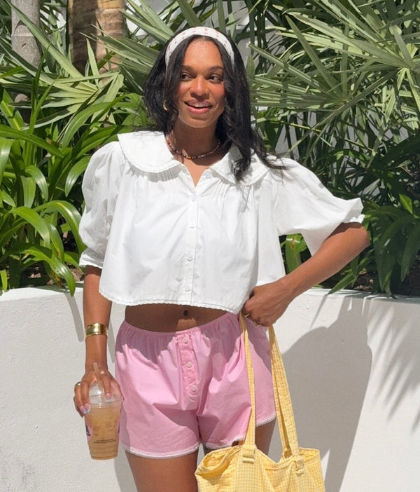 Woman in white blouse and pink shorts standing outdoors with greenery in the background