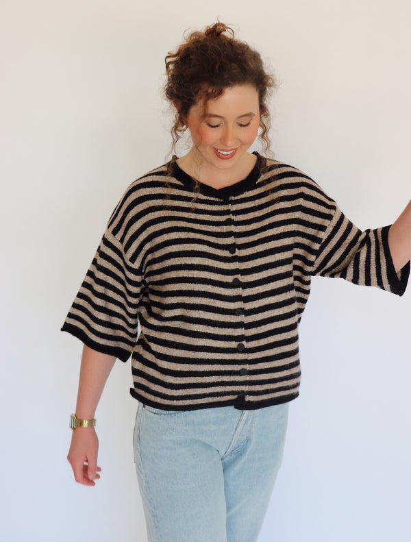 A woman wearing a fuzzy black and beige striped cardigan with three-quarter sleeves and light blue jeans, standing against a plain white wall.