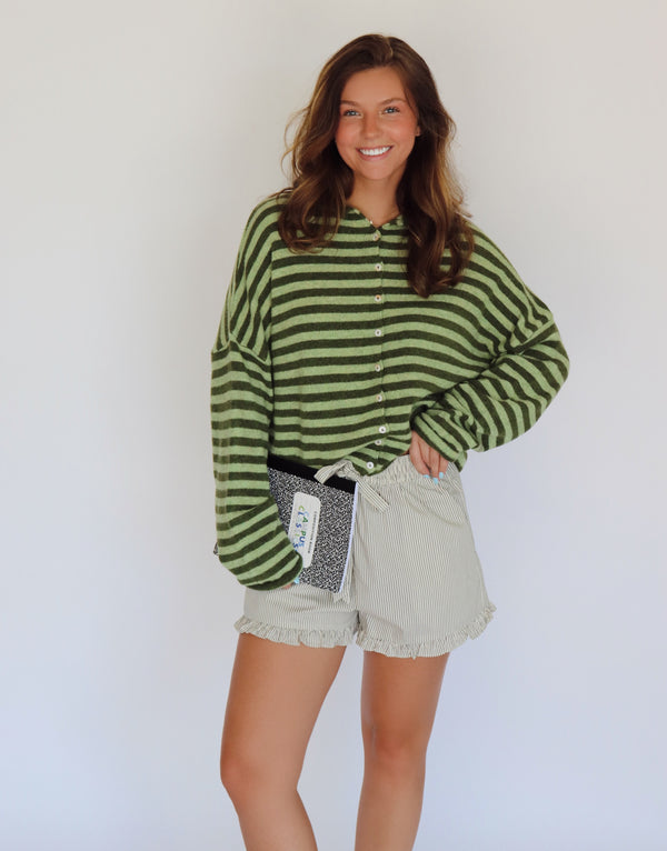 A woman modeling a green and dark green striped long-sleeve button-up cardigan with light beige ruffled shorts, holding a black and white patterned notebook against a plain white background.