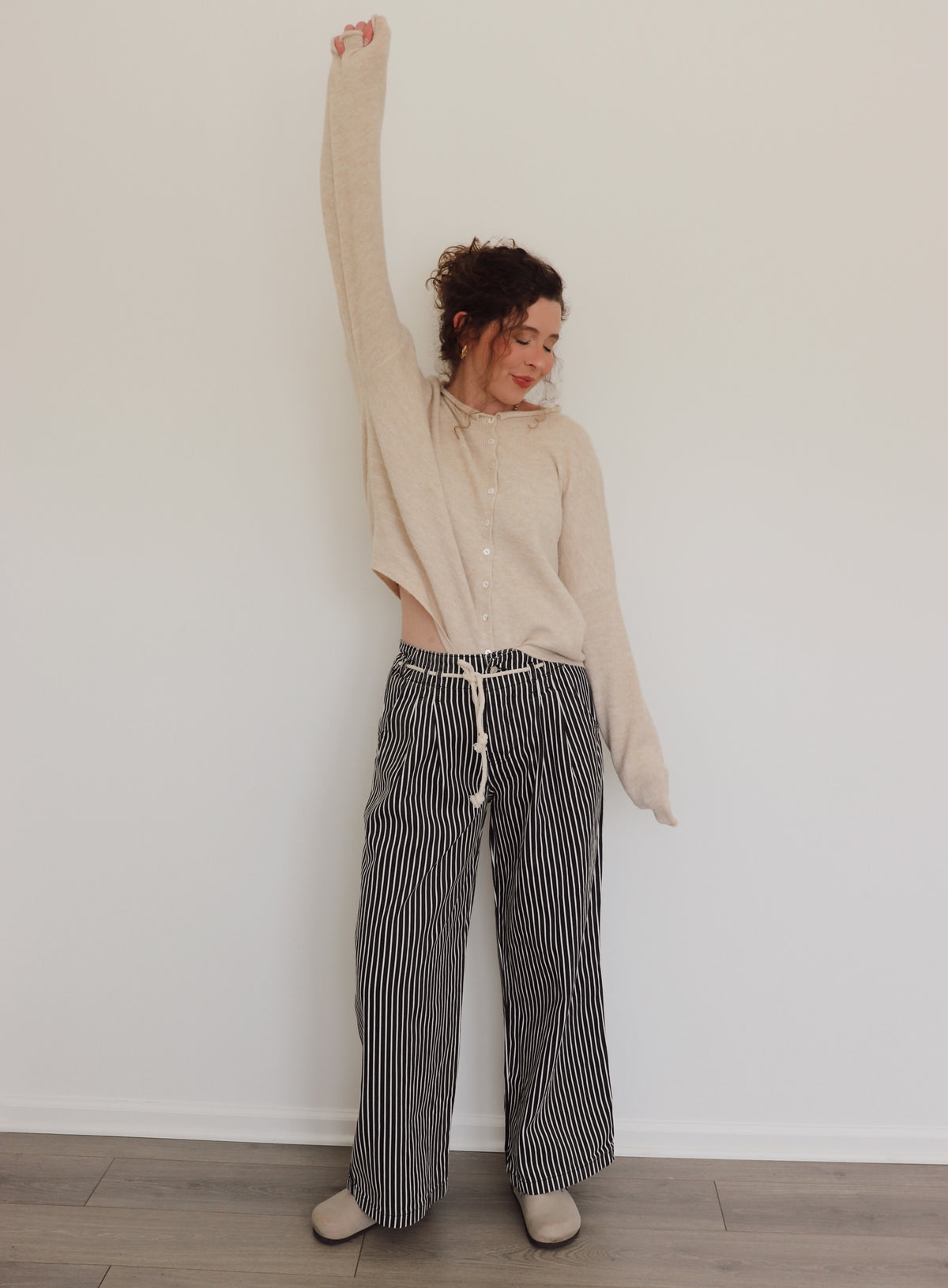 Woman wearing a beige long-sleeve top and black and white striped pants against a plain wall.