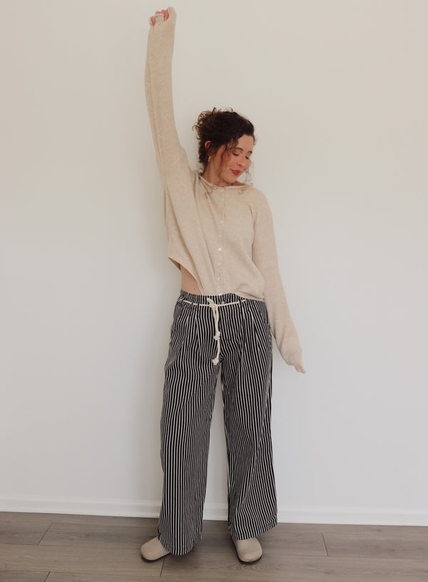 Woman wearing a beige long-sleeve top and black and white striped pants against a plain wall.