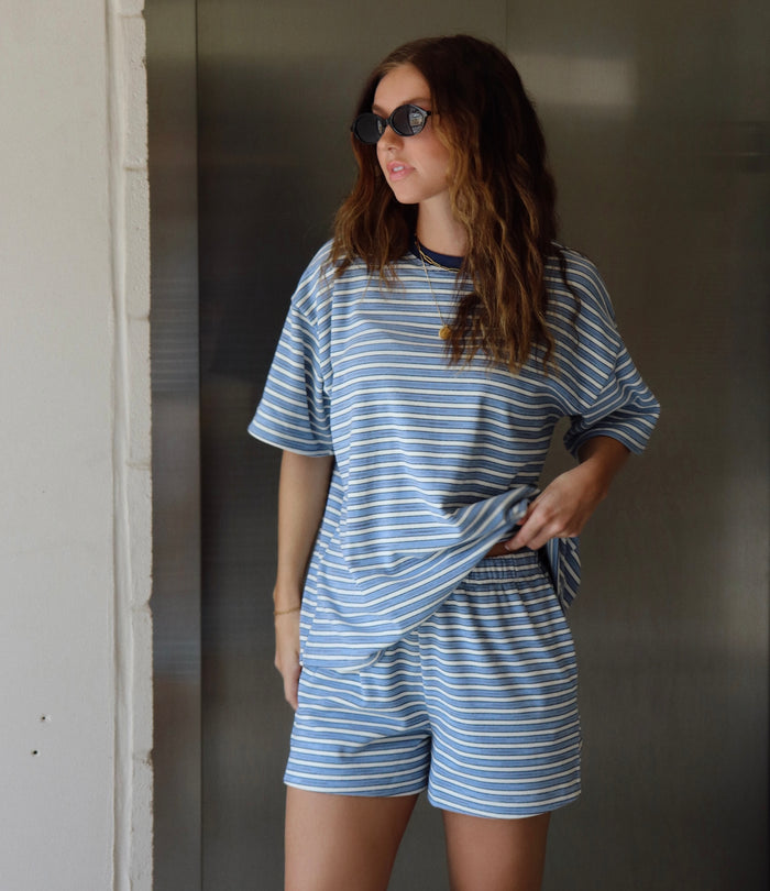 A woman wearing a matching blue and white striped lounge set consisting of an oversized short-sleeve t-shirt and shorts, posing in front of a metallic elevator door.