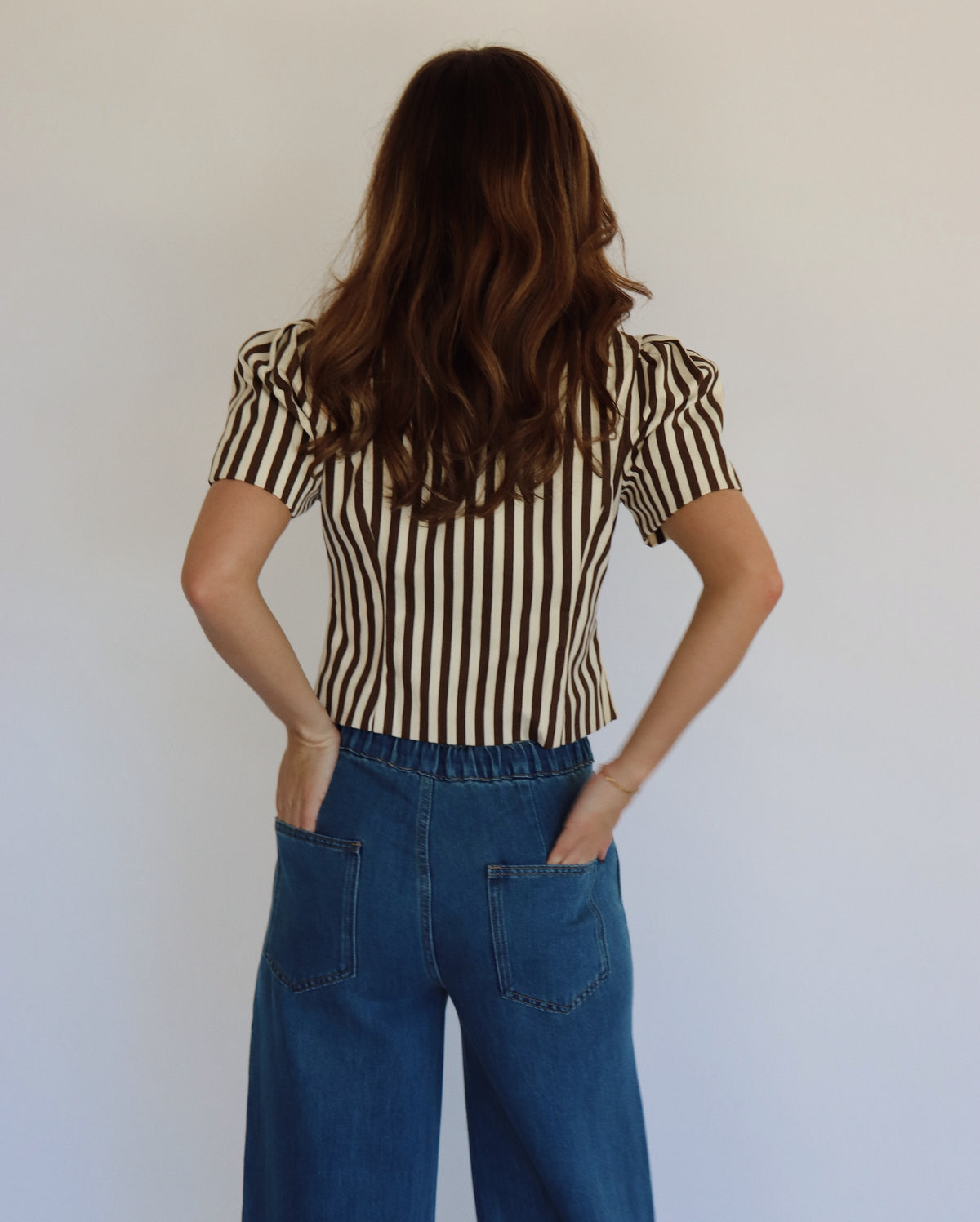 A woman wearing a brown and beige vertically striped short-sleeve button-up top with blue wide-leg jeans, standing against a plain light gray wall.