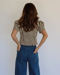 A woman wearing a brown and beige vertically striped short-sleeve button-up top with blue wide-leg jeans, standing against a plain light gray wall.