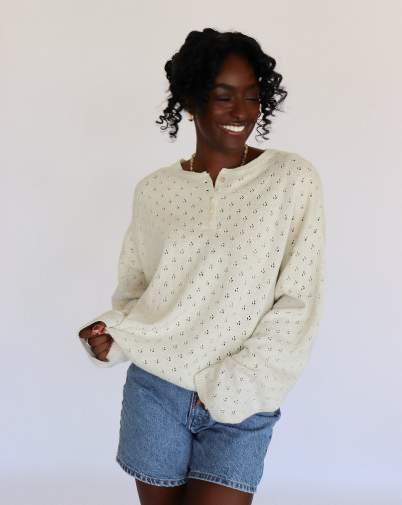 Woman wearing a cream/light gray sweater with small pointelle patterns and denim shorts on a white background