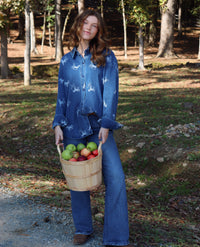A woman stands outdoors in a wooded area holding a wooden basket filled with green and red apples. She is wearing a blue long-sleeve button-up shirt with white horse prints and blue jeans.