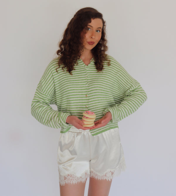 A woman modeling a light green and white striped long-sleeve knit cardigan with white satin shorts, holding a small stack of cookies against a plain light gray background.
