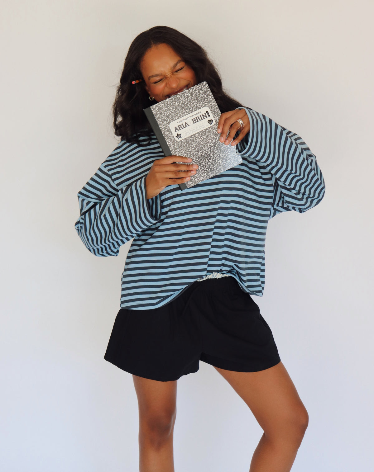 A woman modeling a long-sleeve blue striped top with shorts, holding a black and white notebook bag against a plain white background.