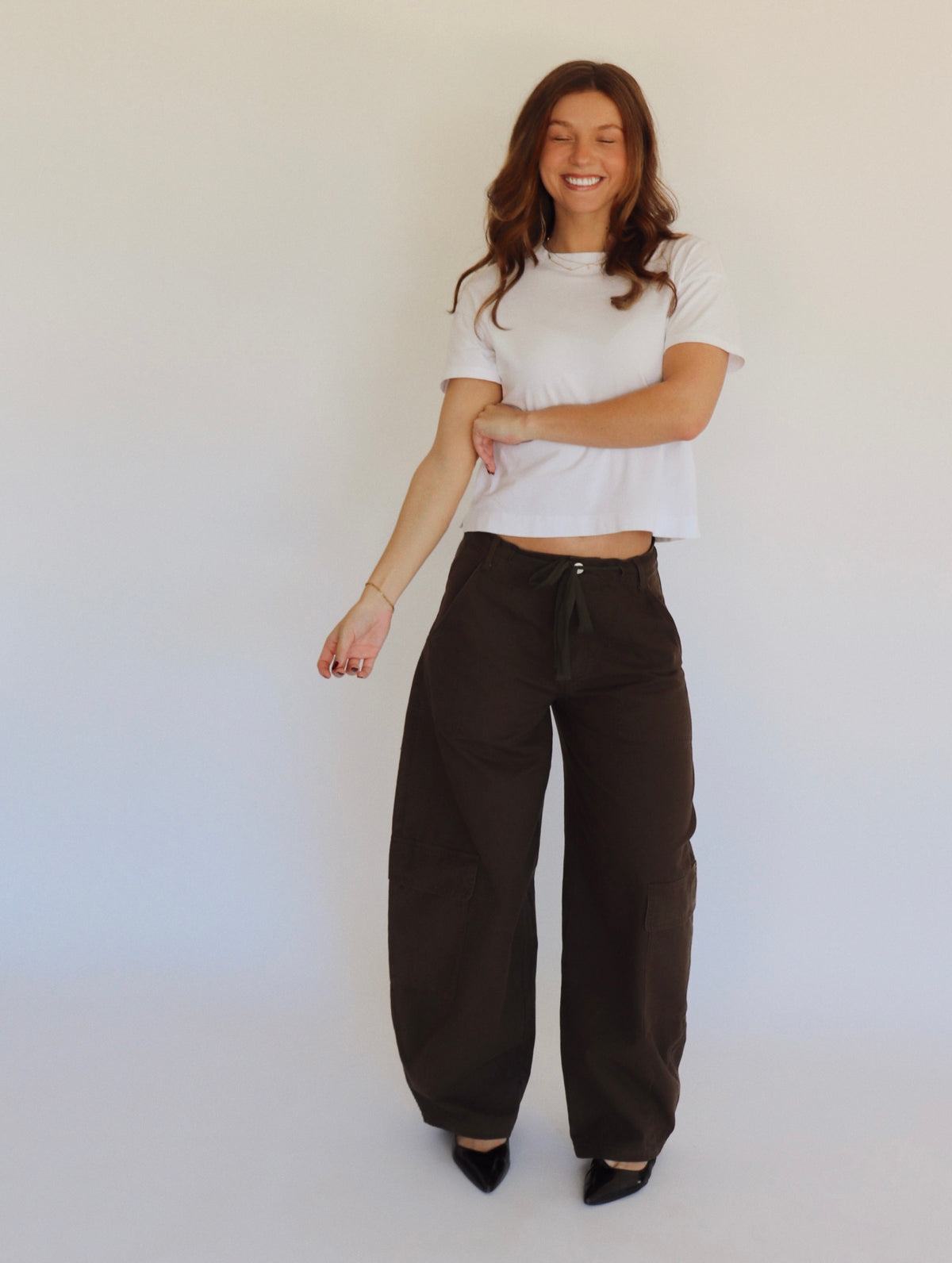 A woman stands against a plain white studio background wearing a white cropped short-sleeve top, loose dark brown wide-leg pants, and black pointed shoes.