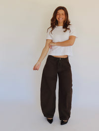 A woman stands against a plain white studio background wearing a white cropped short-sleeve top, loose dark brown wide-leg pants, and black pointed shoes.