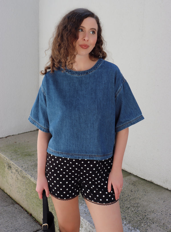 Woman wearing a denim top and polka dot shorts against a beige wall.