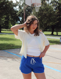 Woman standing on a basketball court wearing a white top and blue shorts.