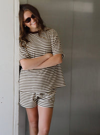 A woman wearing a matching beige and dark brown horizontally striped lounge set, consisting of a short-sleeve top and shorts, posing with her arms crossed against a gray wall.