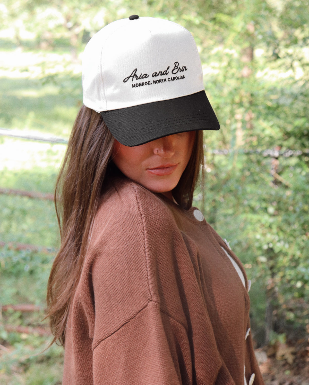 A woman wearing a white and black baseball cap with embroidery on the front, paired with a brown knit cardigan, set against a blurred green outdoor background.