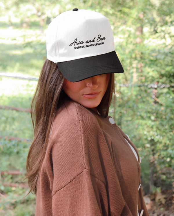 A woman wearing a white and black baseball cap with embroidery on the front, paired with a brown knit cardigan, set against a blurred green outdoor background.