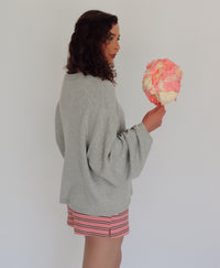 A woman wearing a light gray knit cardigan and pink striped shorts holds a pink cotton candy against a plain white background.
