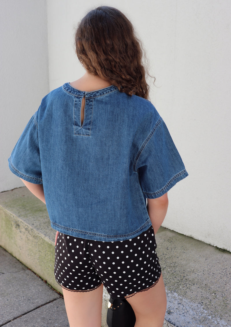 Woman wearing a denim top and polka dot shorts against a beige wall.