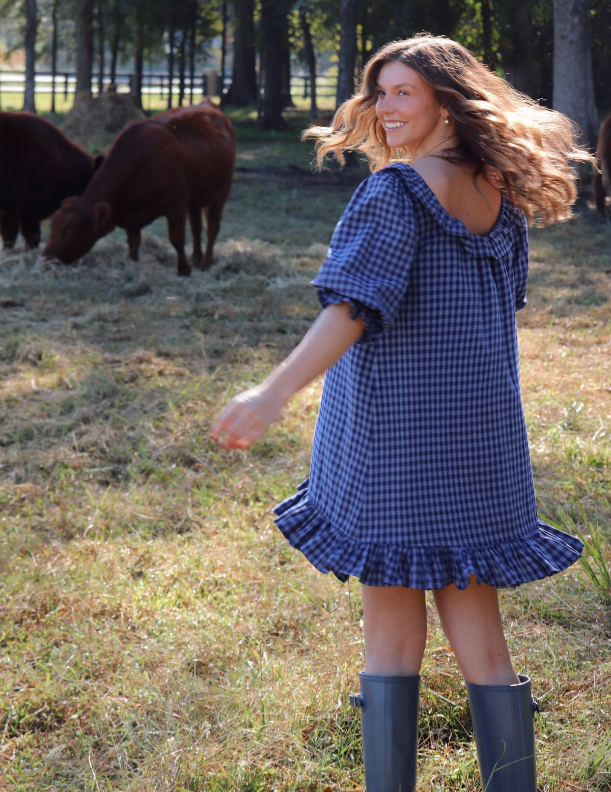 A woman wearing a blue gingham puff-sleeve dress with a ruffled neckline and hem, standing in a grassy field with cows in the background. She is wearing rain boots.