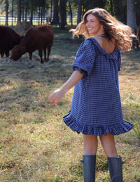 A woman wearing a blue gingham puff-sleeve dress with a ruffled neckline and hem, standing in a grassy field with cows in the background. She is wearing rain boots.