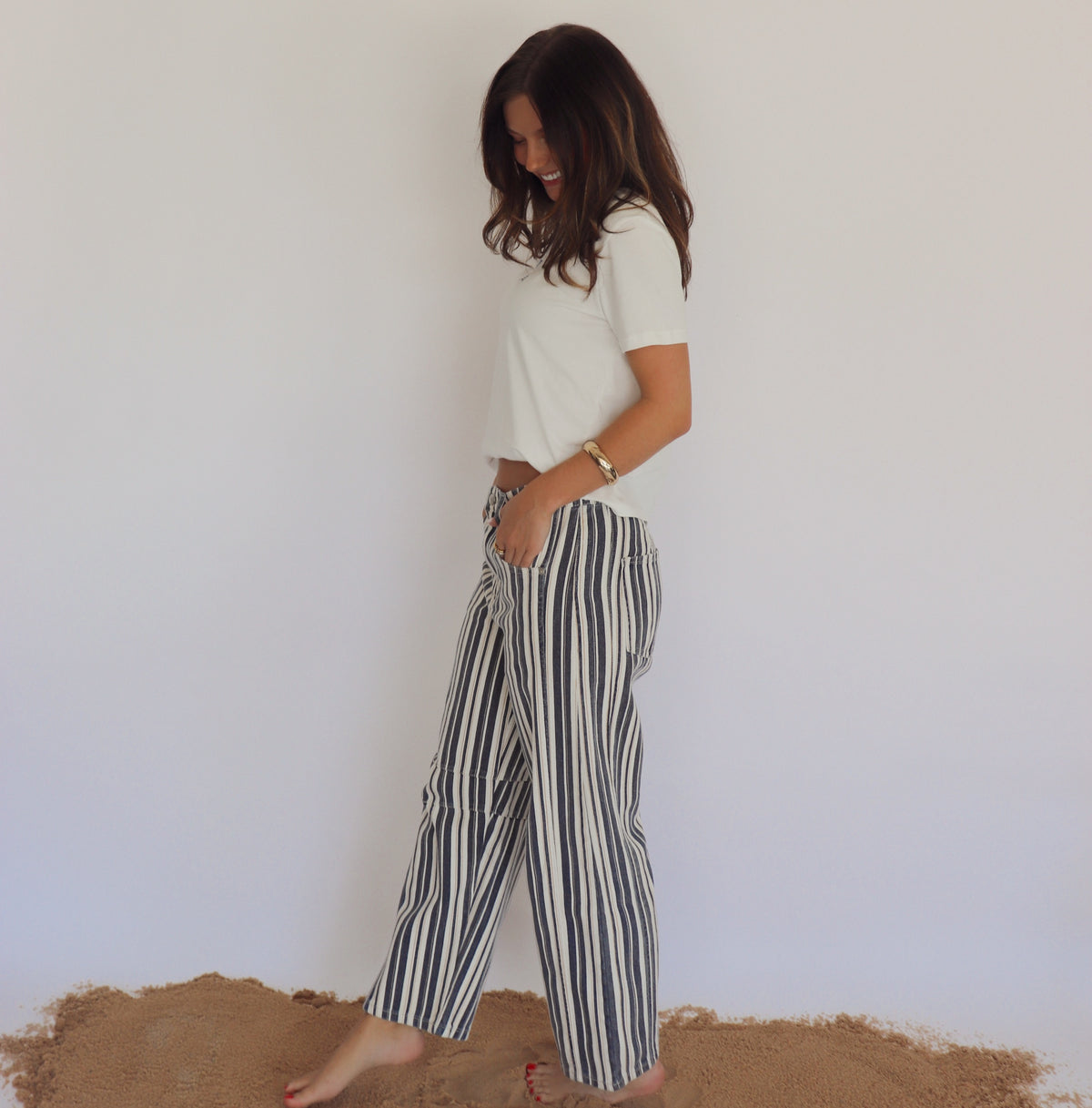 Woman wearing a white top and striped pants standing on a sandy surface with a plain background